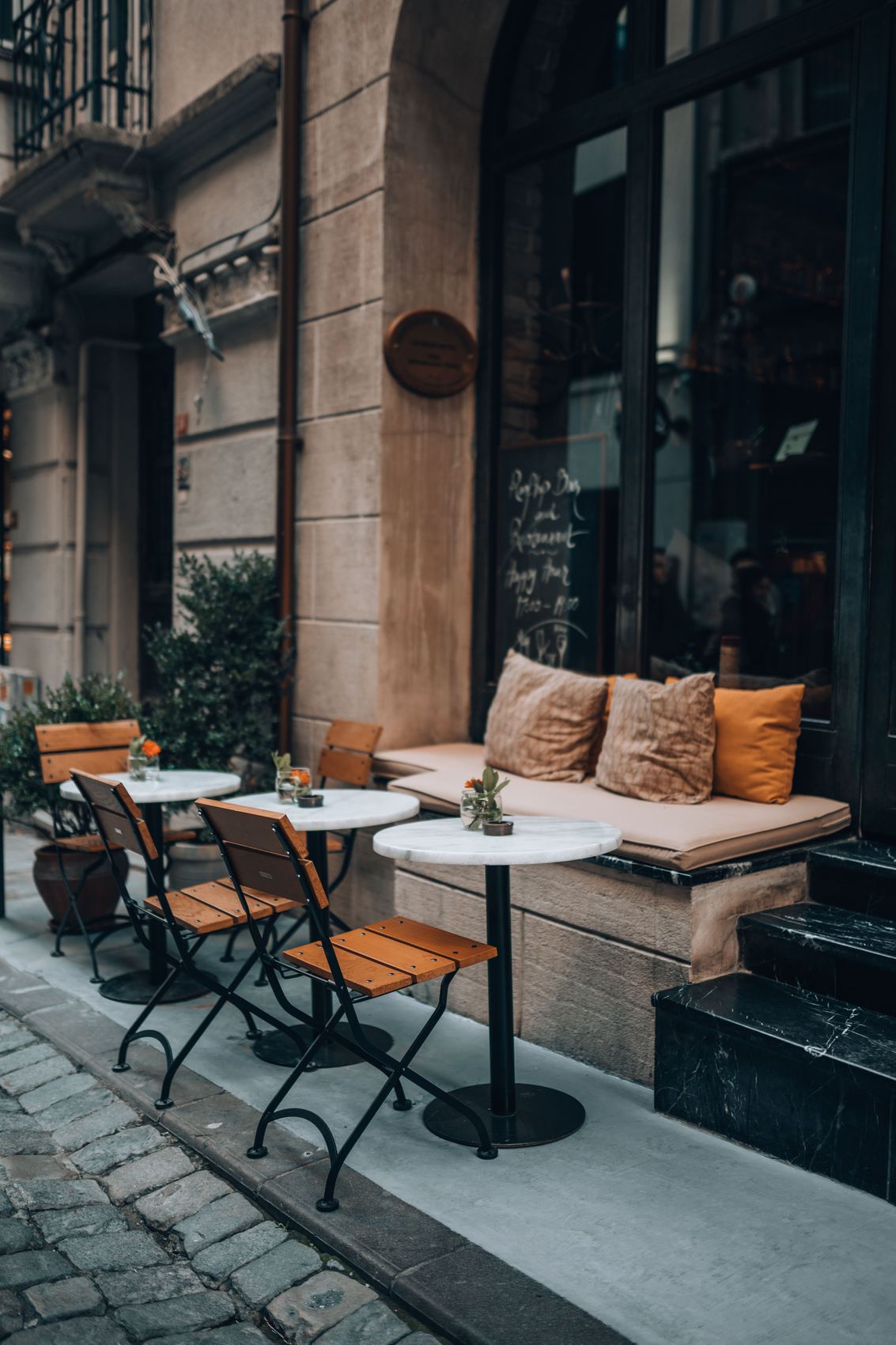 Rustic wooden chairs and small dining tables on an outdoor cafe terrace with potted greenery