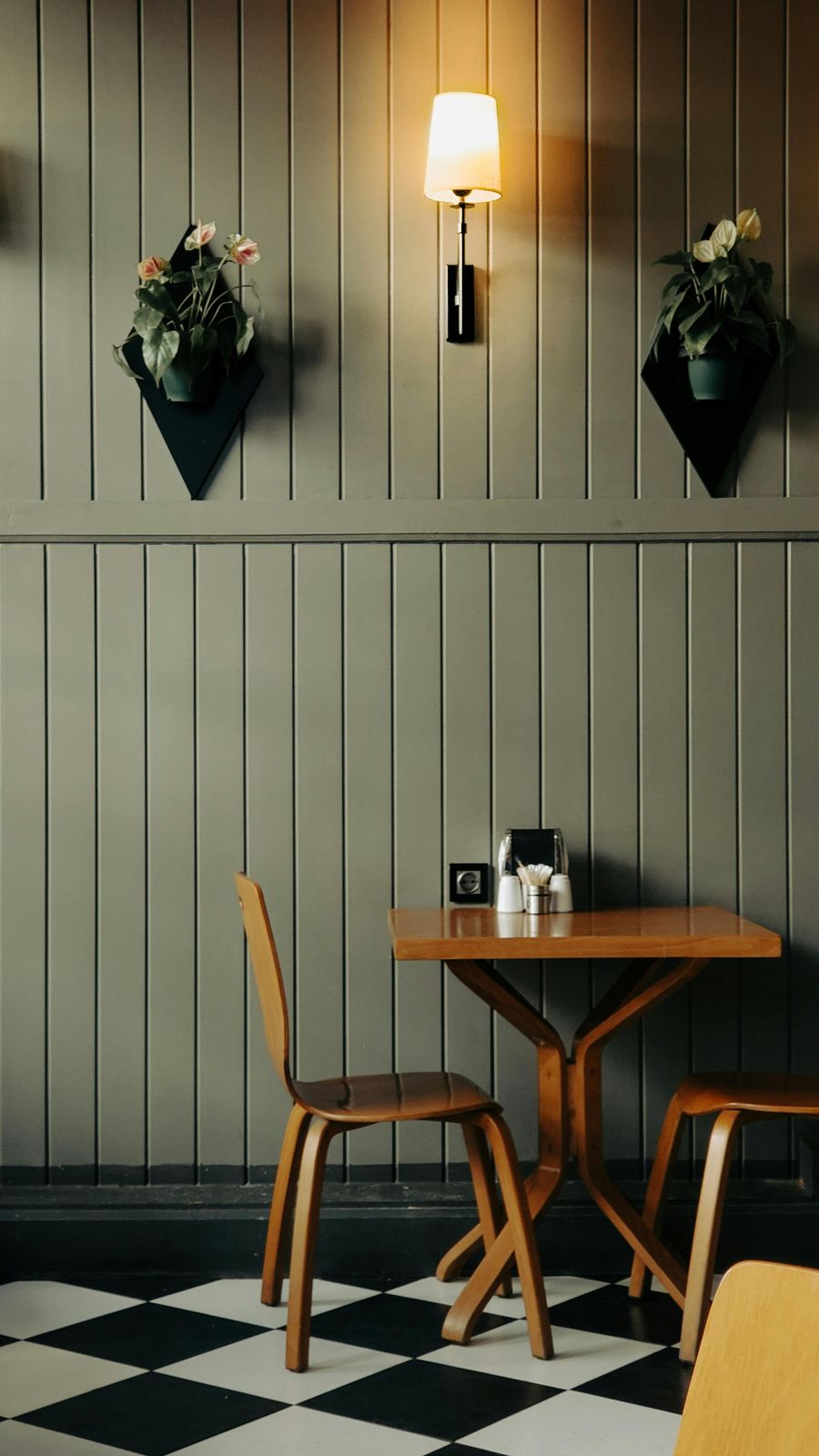 A quiet cafe corner with a wooden table and chairs against a panelled wall, lit by a single warm wall lamp