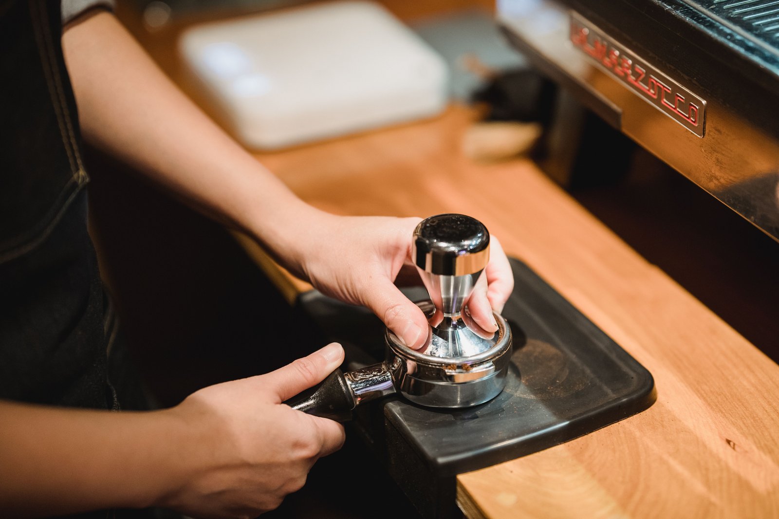 Close-up of a barista's hands tamping espresso grounds into a portafilter at an espresso machine, no face in frame