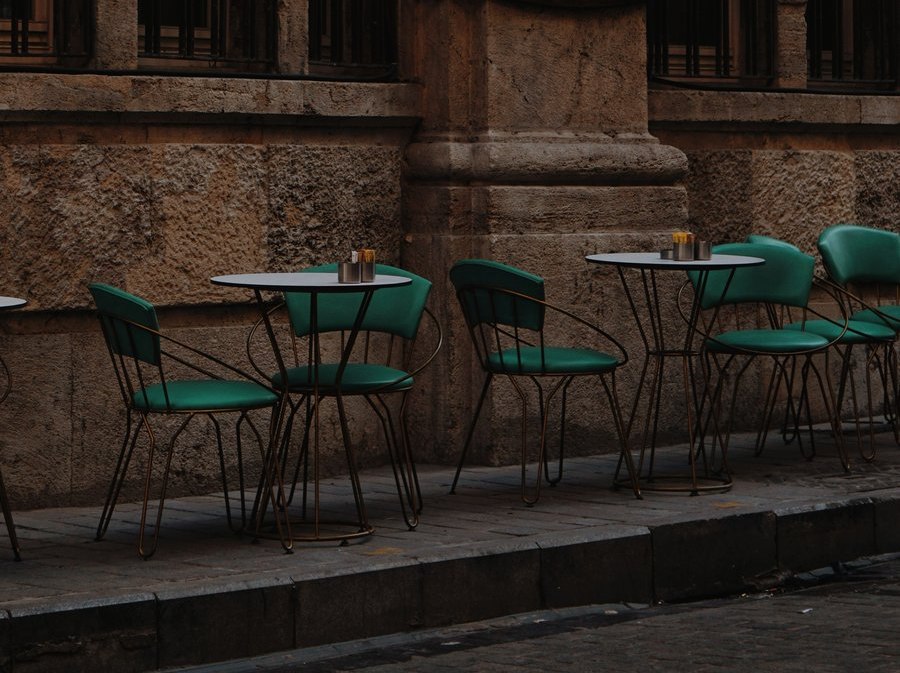 Small round cafe tables with green chairs set along a stone-paved sidewalk against a weathered stone building facade